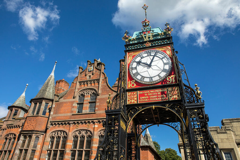 A large ornate clock, the Eastgate Clock, stands on a decorative iron structure in front of a historic red brick building under a blue sky with scattered clouds.