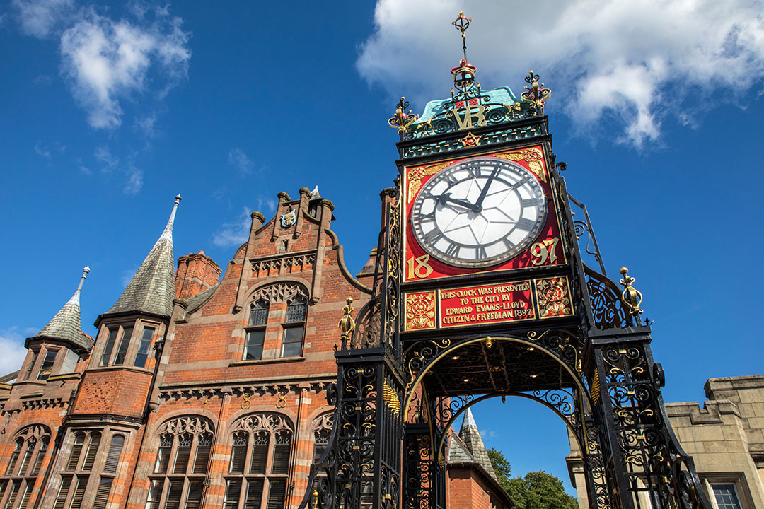 A large ornate clock, the Eastgate Clock, stands on a decorative iron structure in front of a historic red brick building under a blue sky with scattered clouds.