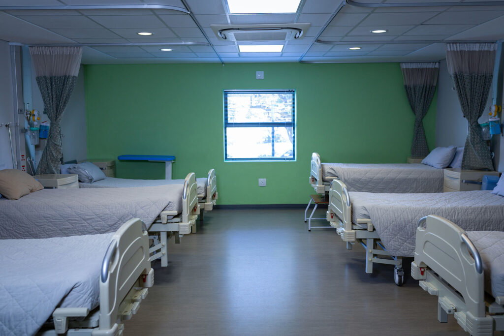 You are in a bright hospital ward with six empty beds separated by curtains. Each bed faces a window on a green wall, and medical equipment is mounted on the walls behind the beds. Overhead lights provide ample lighting.