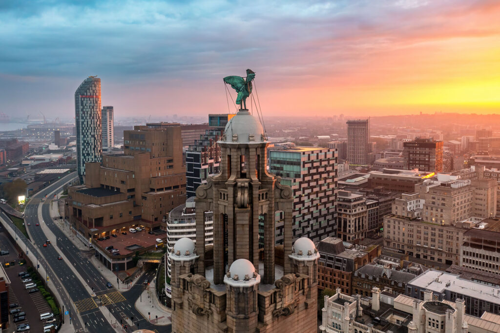 Aerial sunset view of Liverpool, highlighting the Royal Liver Building with its famous Liver Bird statue, surrounded by a mix of modern glass towers and historic brick buildings for a rich cityscape experience.