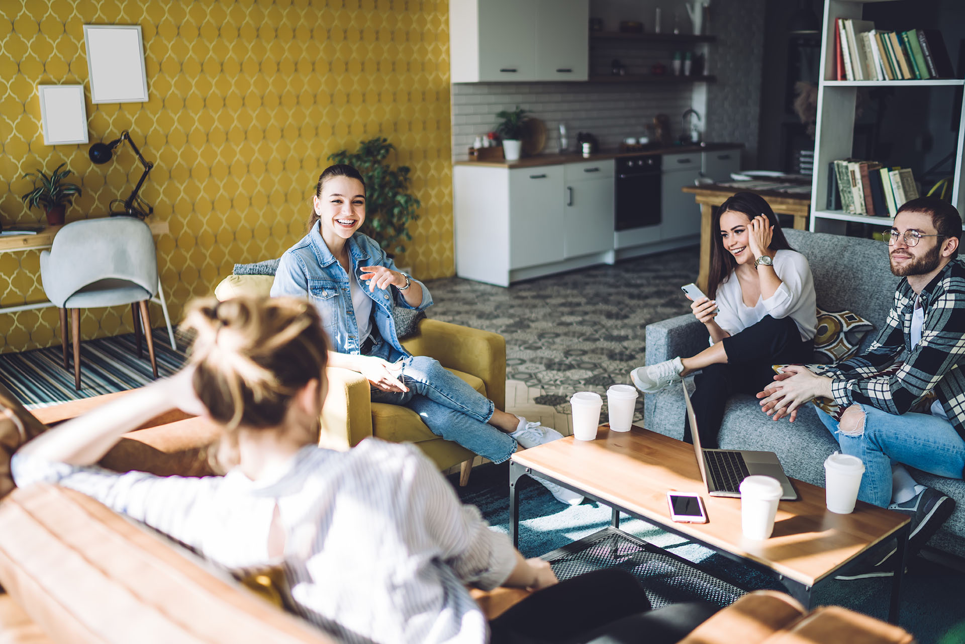 You’re in a cosy living room with four people chatting and smiling. Tables hold coffee cups, a laptop, and a mobile phone. Behind them, there’s a friendly yellow wall, a kitchen area, green plants, and tidy shelves.