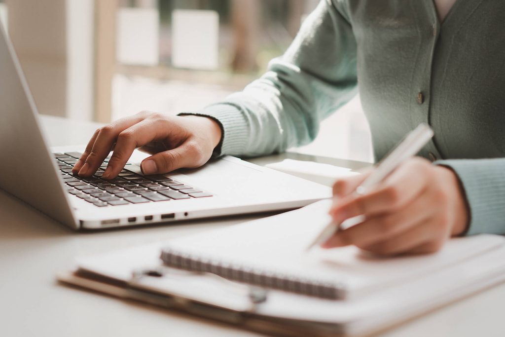 You see someone in a green jumper multitasking at a desk, typing on a laptop with one hand and writing in a spiral notebook with the other, balancing digital and handwritten tasks to stay organised.