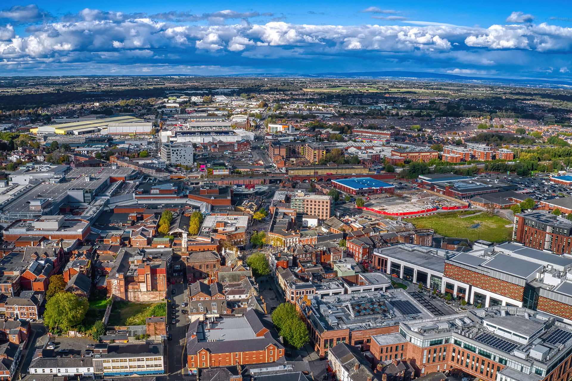 Looking for removals Cheshire services? The image shows an urban area from above, with tightly packed buildings, industrial zones, leafy green spaces, and connecting roads all under a blue sky dotted with clouds.