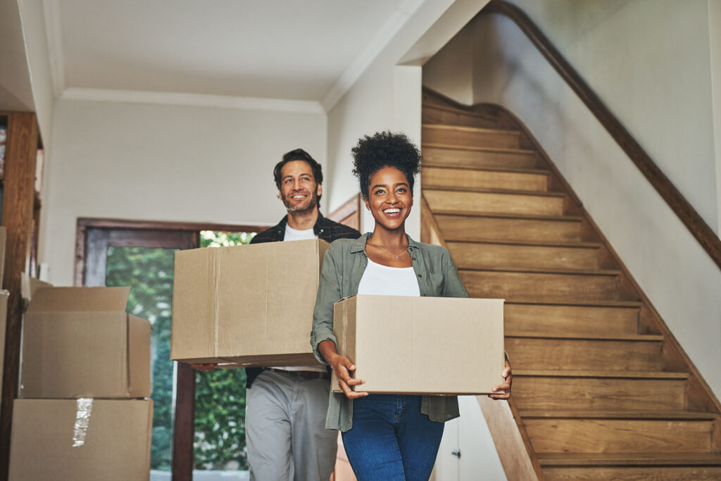 Smiling removals workers with cardboard boxes step into a bright home, demonstrating a stress-free moving process. A well-lit staircase in the background aids navigation, helping you visualise an accessible move-in experience.