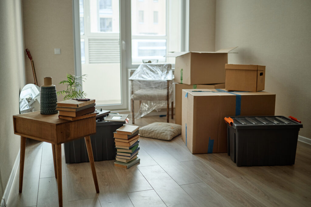 You are viewing a room filled with packed moving boxes, plastic storage containers, a table stacked with books, a single chair, and a potted plant placed near the window—suggesting an active moving process.