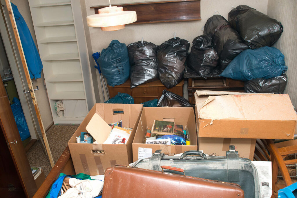 A room filled with stacked rubbish bags on furniture, open cardboard boxes containing assorted belongings, and a suitcase positioned in the foreground, making navigation and identification of items challenging.