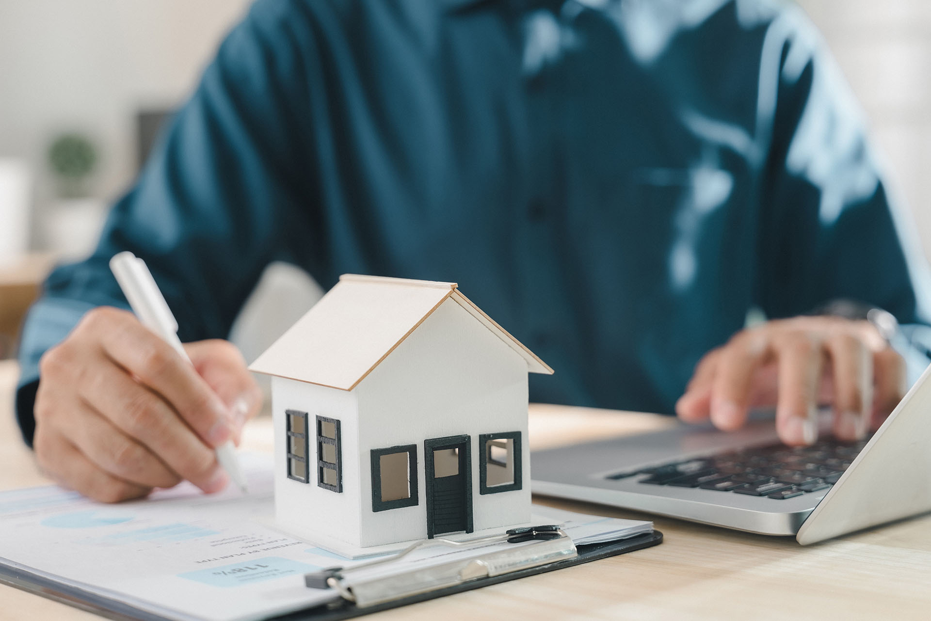 A person wearing a blue shirt sits at a desk with a laptop and paperwork beside a model house, organising contacts to notify about their move—helpful for customers handling property or estate changes.