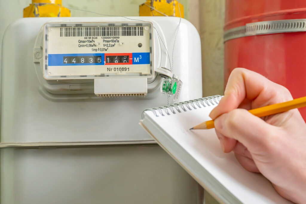 You’re recording numbers from a household gas meter with a blue and red scale, holding a pencil and notepad during your first week in your new home. The image focuses on careful tracking for your records.