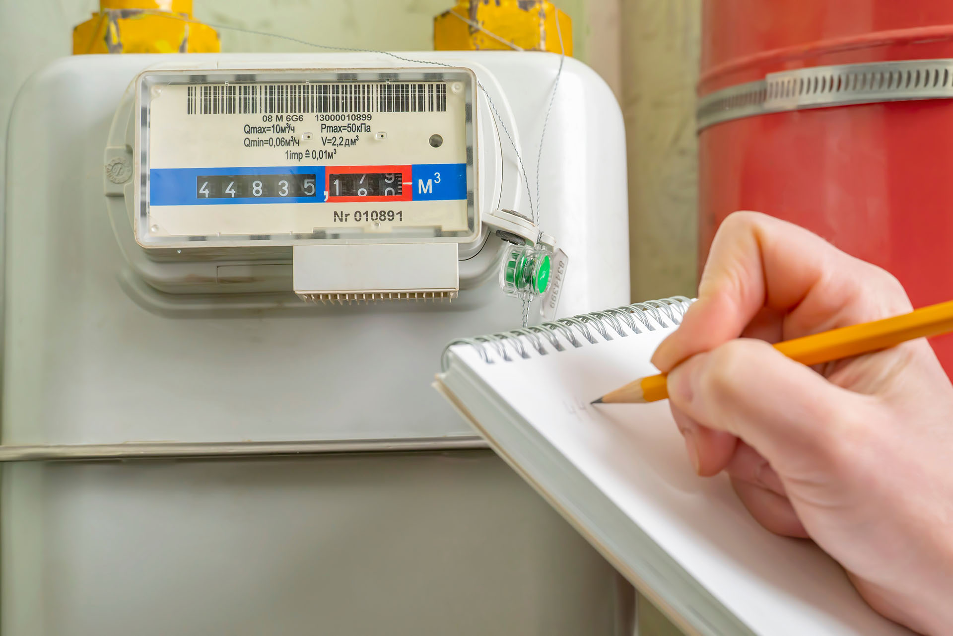 You’re recording numbers from a household gas meter with a blue and red scale, holding a pencil and notepad during your first week in your new home. The image focuses on careful tracking for your records.