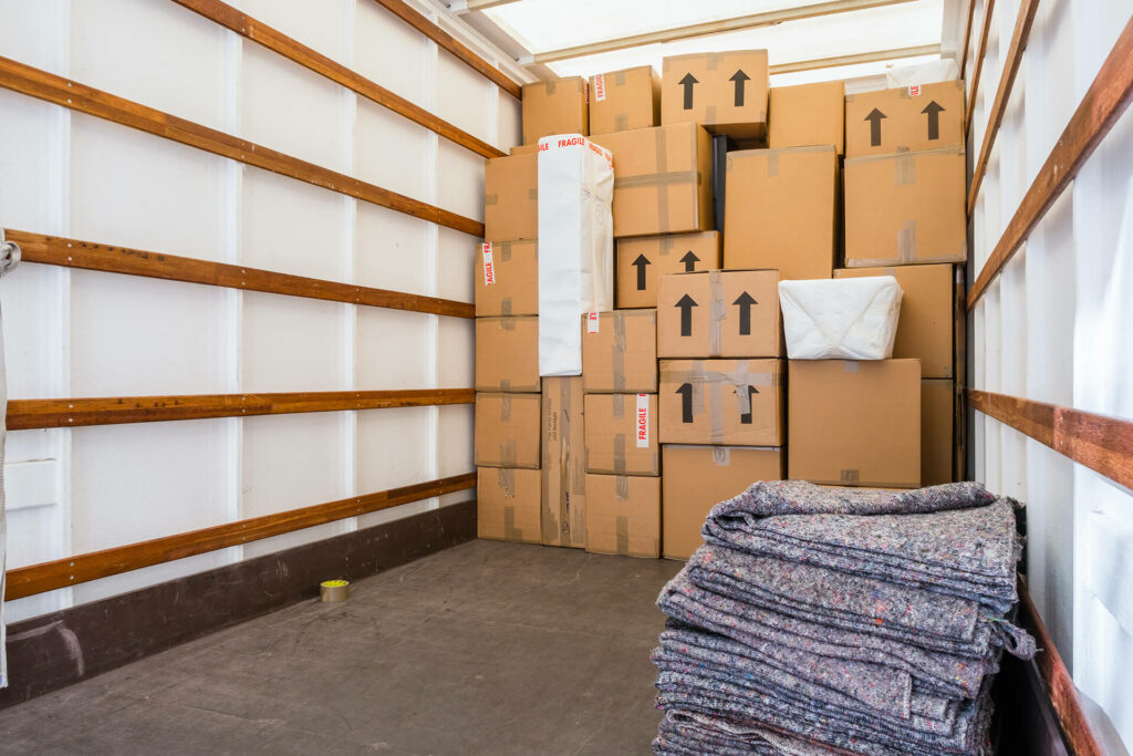 You are looking into a moving lorry with neatly organised cardboard boxes stacked along the wall. In the front, there’s a folded pile of removal blankets on the floor, ready to protect your belongings.