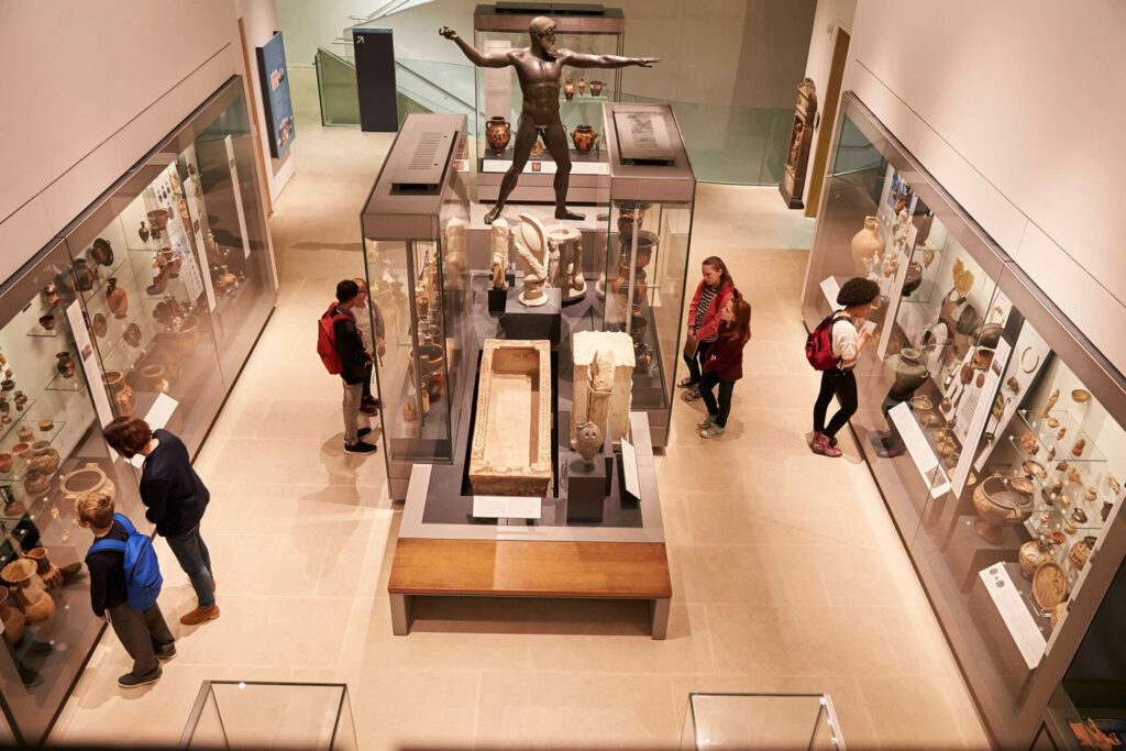 You can explore ancient artefacts and sculptures displayed in glass cases at the museum, featuring a central bronze statue with outstretched arms and a stone sarcophagus among the exhibits.