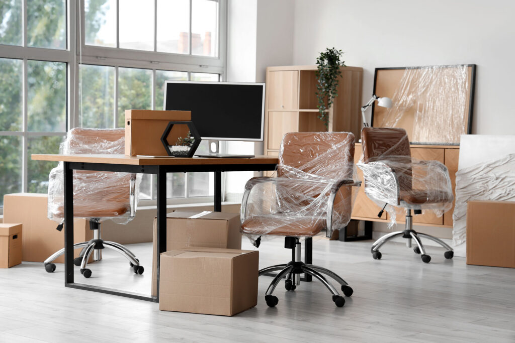 Bright, spacious office ready for you, with new swivel chairs still in plastic, a desk featuring a monitor, unopened cardboard boxes, and shelves beside large windows letting in plenty of natural light.