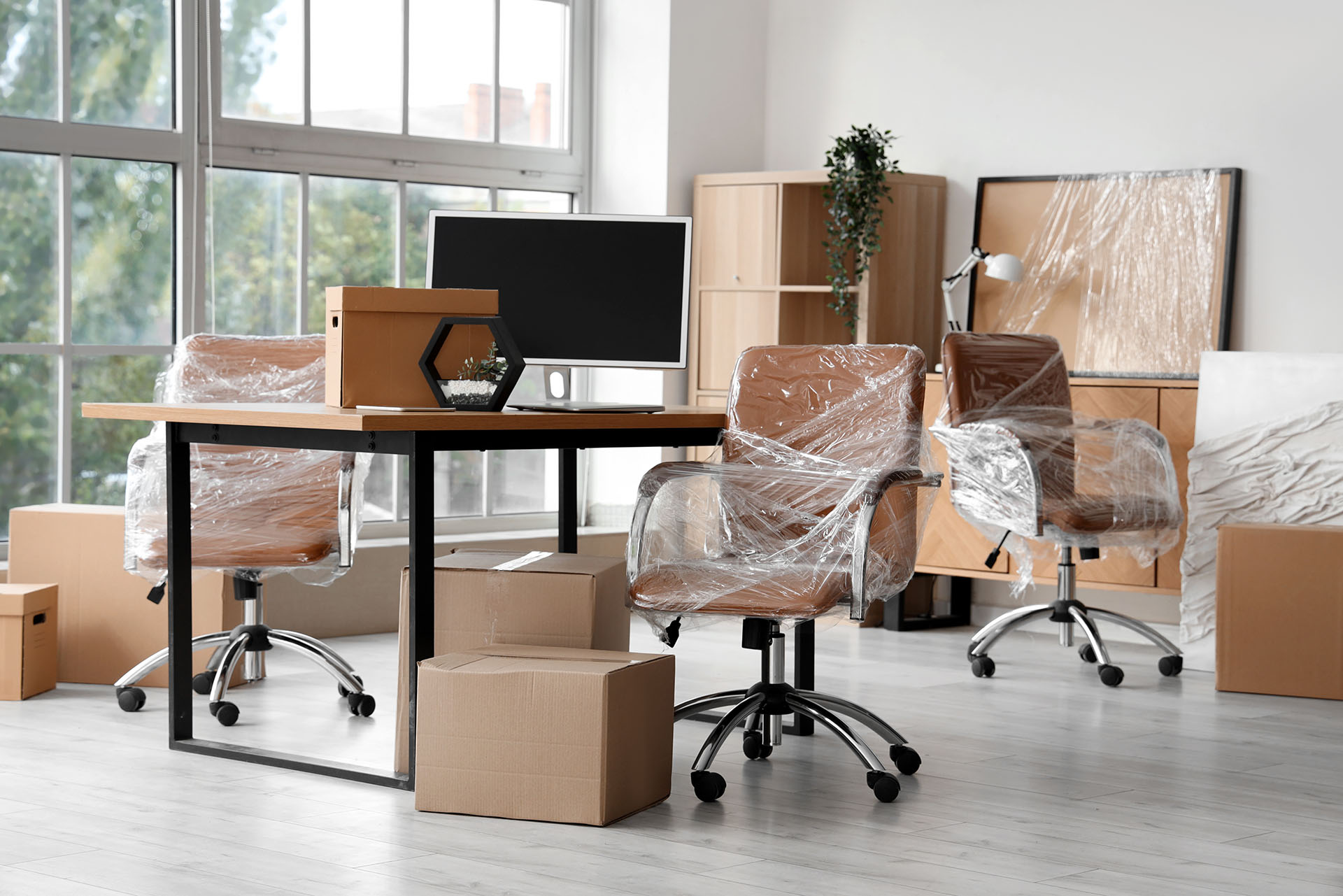 Bright, spacious office ready for you, with new swivel chairs still in plastic, a desk featuring a monitor, unopened cardboard boxes, and shelves beside large windows letting in plenty of natural light.