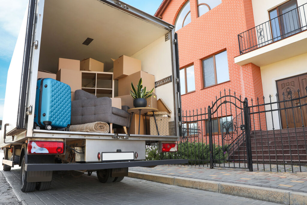 A removal lorry waits outside your new brick house, filled with boxes, a blue suitcase, sofa, plant, and other household belongings—ready for you to settle into your next chapter.