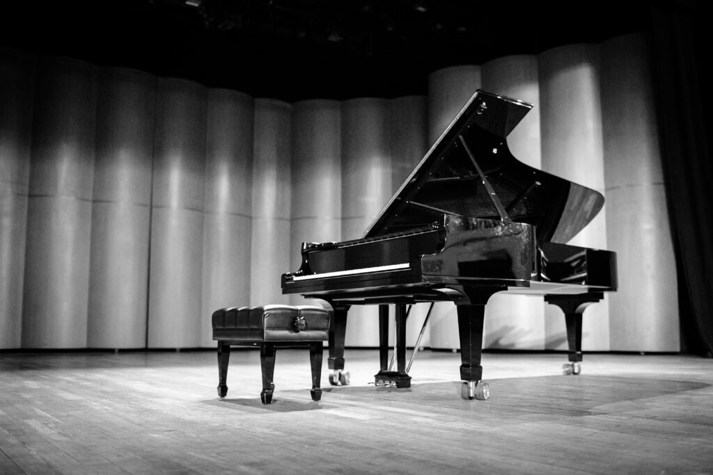 You’ll find a grand piano with a stool on a polished wooden stage, set before a smoothly curved backdrop. Overhead stage lights create gentle shadows to help you imagine the concert setting.