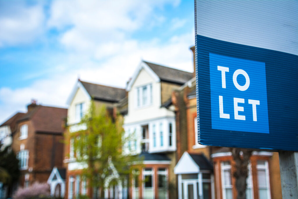 A prominent “To Let” sign stands at the front of a row of classic British houses, ideal for prospective tenants. Lush trees and a clear blue sky create a welcoming, serene neighbourhood setting.