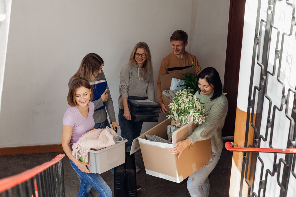Five individuals are seen carrying boxes and household belongings up a stairwell, likely moving into a new residence. The scene shows teamwork as they navigate the stairs with various items in hand.