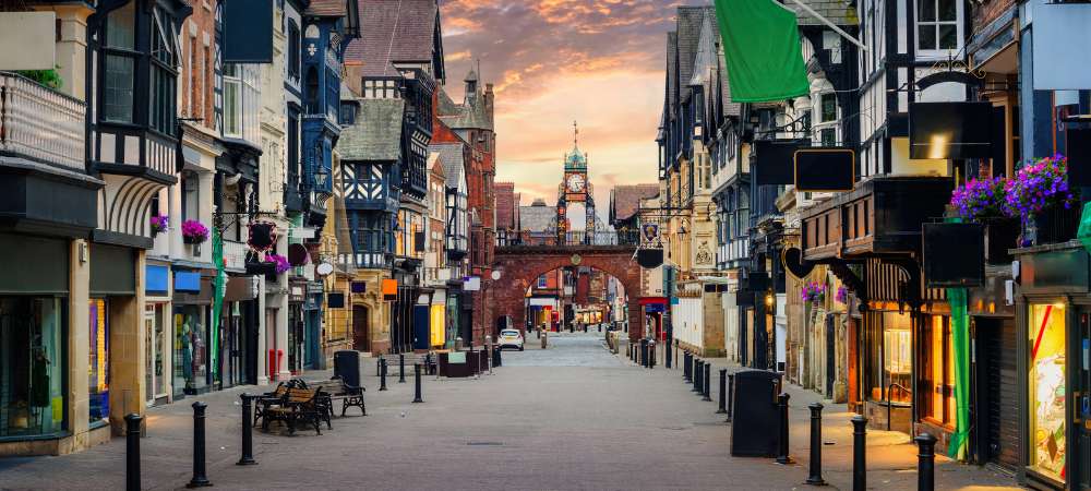 As you move along a street featuring timber-framed buildings and shops, you'll approach an archway crowned by a clock tower, showcasing the historic charm typical of removals Cheshire routes.