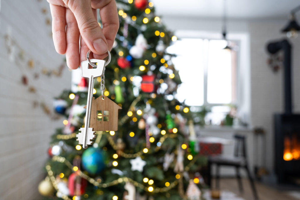 A hand holds a key featuring a small, wooden, house-shaped keyring, positioned in front of a festive, decorated Christmas tree.