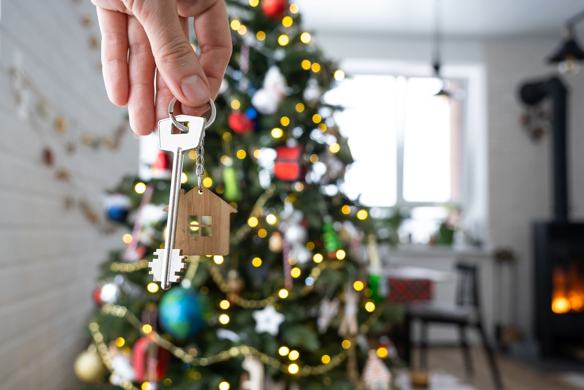 A hand holds a key featuring a small, wooden, house-shaped keyring, positioned in front of a festive, decorated Christmas tree.