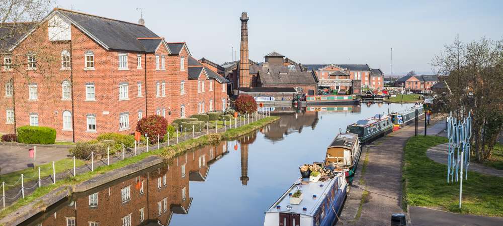 Enjoy a serene view of a sunlit canal in Cheshire, where colourful narrowboats, red brick buildings, and a tall old chimney are mirrored perfectly in the calm water—a peaceful sight near our removals service.