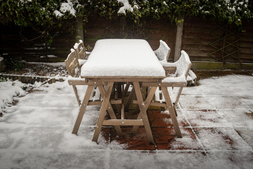 A wooden patio dining set sits outdoors, blanketed in a layer of snow during winter storage.