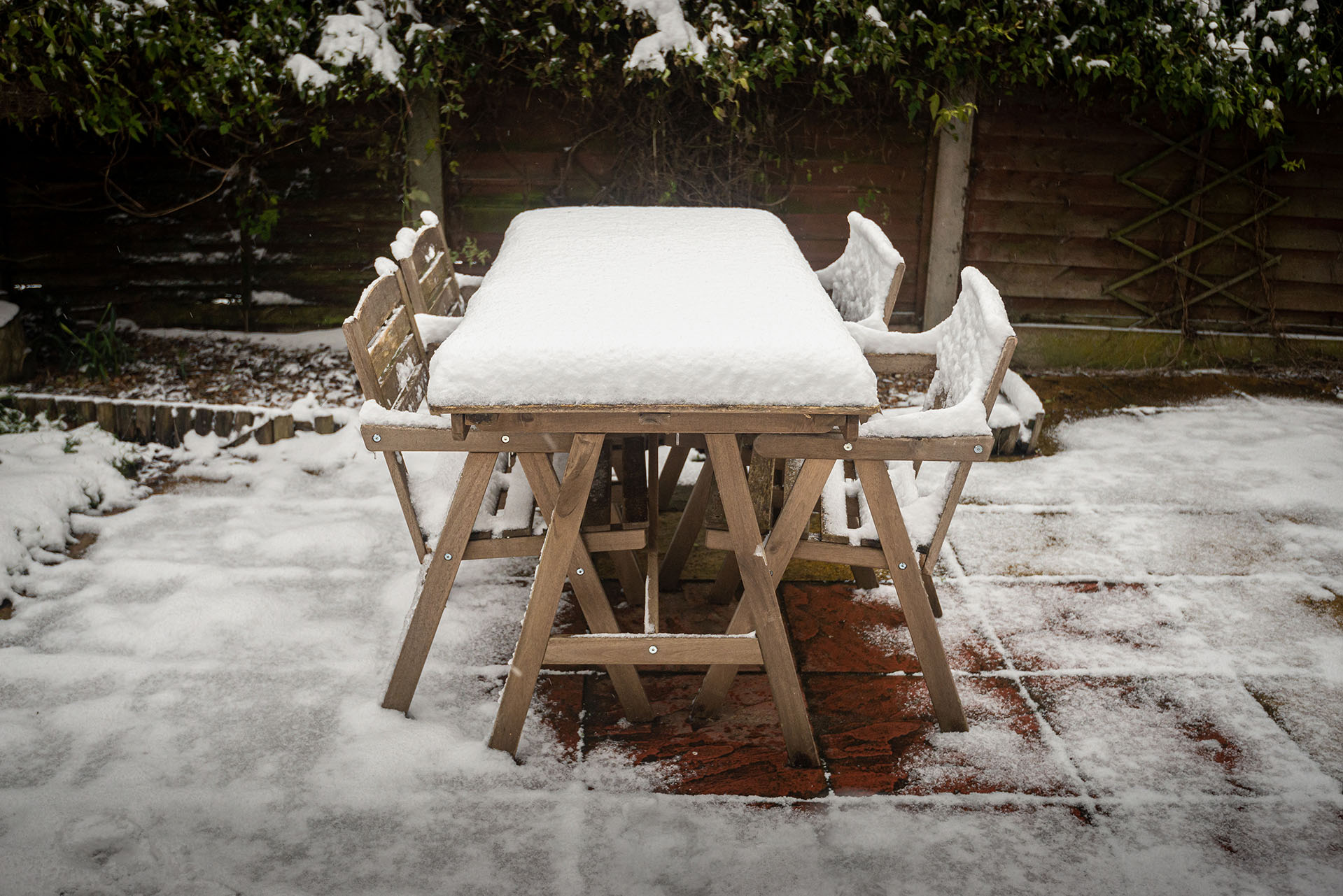 A wooden patio dining set sits outdoors, blanketed in a layer of snow during winter storage.