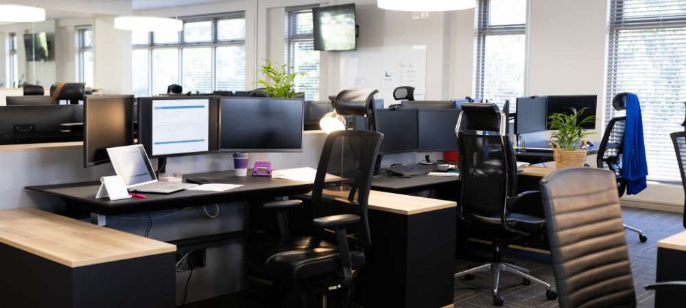 Bright, modern open-plan office with spacious workstations, computer monitors, and greenery. Large windows with blinds create a welcoming environment ideal for your team or Cheshire business removals needing an organised setting.