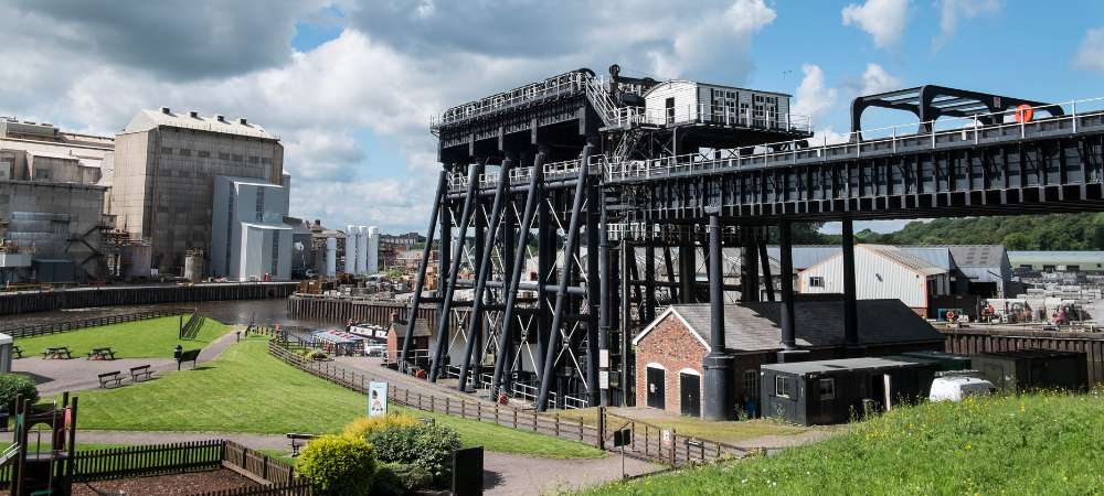 A large black metal boat lift stretches across a canal by industrial buildings, a grassy park, and a small brick building in Cheshire, all under a partly cloudy sky—showcasing local engineering and shipping activity.