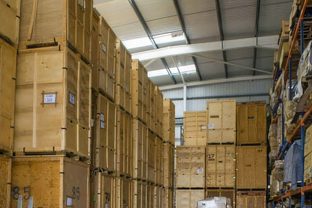 You’re viewing neatly stacked large wooden crates, each clearly labelled and organised in rows within a spacious warehouse. Metal roofing covers the space, and additional metal shelving is visible on the right.