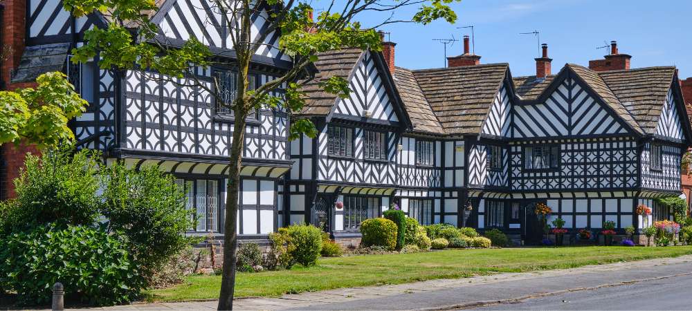 You’ll find a row of striking black and white Tudor-style houses with gabled roofs, neat lawns, and bright flowerpots under a blue sky—a welcoming glimpse into Cheshire neighbourhoods during your move.